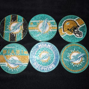 May include: Six circular coasters decorated with colorful rhinestones featuring Miami Dolphins logos and football helmet designs in shades of teal, orange, white, and black. Each coaster has a distinct pattern, including stripes and text reading MIAMI DOLPHINS.