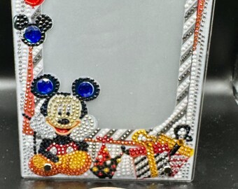 May include: A rectangular photo frame decorated with rhinestones, featuring Mickey Mouse and gifts. The frame has red, blue, and yellow accents. A wooden disc with the text "Creations by Day" is in the foreground.