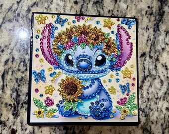 May include: Square artwork featuring a blue cartoon character with large eyes, adorned with a floral crown and holding a sunflower. The image is embellished with colorful gems and butterflies, set against a light background.