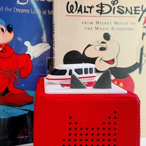 Op de afbeelding: Een rode kubusvormig speelgoed met een witte bovenkant, met een speelgoedtrein en twee grijze haaienvinnen. Achter het speelgoed staan twee Disney-boeken, één met de titel "Imagine" en de andere "The Art of Walt Disney."