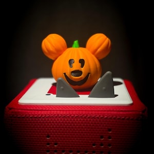 May include: An orange pumpkin-shaped figure with round ears and a black carved face sits atop a red square speaker. The speaker features a white top panel with two small gray triangular accents positioned in front of the pumpkin figure.