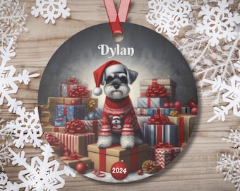 May include: A round Christmas ornament featuring a gray and white miniature schnauzer dog wearing a red Santa hat and a red and white sweater. The dog is surrounded by wrapped presents with red and white bows. The ornament has the name "Dylan" and the year "2024" printed on it.