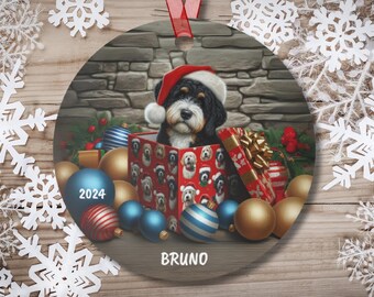 May include: A round Christmas ornament featuring a dog wearing a Santa hat, inside a gift box. The box is surrounded by Christmas ornaments and gifts. The year "2024" is displayed on a blue ornament, and the name "BRUNO" is at the bottom.