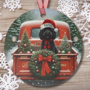 May include: Round Christmas ornament featuring a black dog wearing a Santa hat and a red and black plaid scarf, sitting in the back of a red truck. The truck bed is decorated with a wreath and Christmas trees. The text "RUFUS" is on the hat.