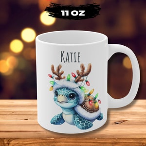 May include: White ceramic mug with a festive turtle design. The turtle wears a reindeer antler hat with colorful Christmas lights. The name "KATIE" is printed above the turtle. The mug is labeled "11 OZ".
