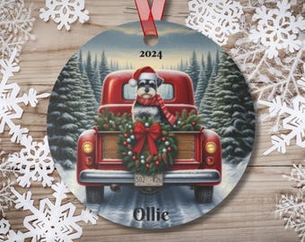 May include: A red vintage pickup truck with a Christmas wreath in the bed. A small gray dog wearing a Santa hat is sitting in the bed of the truck. The truck is driving through a snowy forest. The ornament says "2024" and "Ollie".