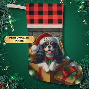 May include: A personalized Christmas stocking with a red and black buffalo plaid cuff. The stocking features a Cavalier King Charles Spaniel dog wearing a Santa hat and peeking out of a gift box. The dog's name, "Coco", is written on the hat.