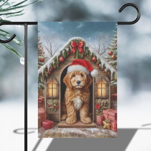 May include: A Christmas-themed garden flag featuring a golden retriever puppy wearing a Santa hat, peeking out of a wooden doghouse decorated with lights, snow, and a red bow. The doghouse is surrounded by presents and a snowy winter scene.