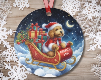 May include: A round Christmas ornament featuring a cartoon dog wearing a Santa hat and riding in a red sleigh filled with presents. The dog is brown and white and has a red bow tie. The sleigh is flying through the air with a crescent moon and stars in the background. The ornament has the text "Charlie 2024" on the side.