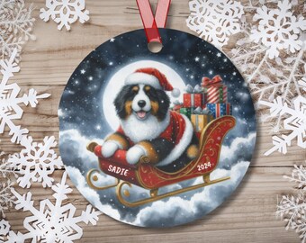 May include: A round Christmas ornament featuring a Bernese Mountain Dog dressed as Santa Claus, pulling a red sleigh filled with presents. The dog is sitting in the sleigh, looking up with a happy expression. The ornament has a blue background with white clouds and stars. The text "SADIE" is written on the sleigh and "2024" is written on the side.