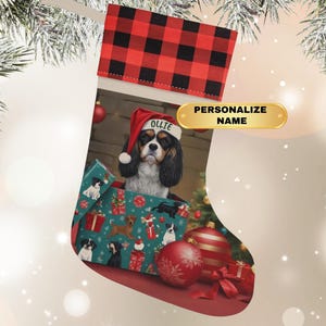 May include: A Christmas stocking with a red and black buffalo plaid cuff. The stocking features a Cavalier King Charles Spaniel wearing a Santa hat with the name "Ollie". The stocking is decorated with Christmas gifts and ornaments.