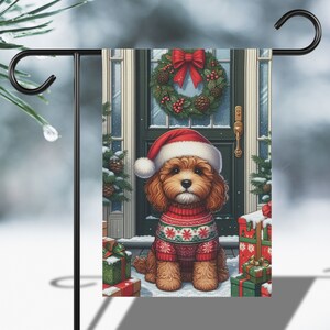 May include: A garden flag featuring a cute brown dog wearing a Santa hat and a red Christmas sweater. The dog sits in front of a decorated door with a wreath, surrounded by wrapped gifts and snow. The flag is mounted on a black metal stand.