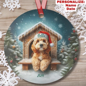 May include: A Christmas ornament featuring a dog wearing a Santa hat, sitting in a doghouse with snow falling around it. The ornament is decorated with a green 2024 and the name "Bella" on the doghouse.
