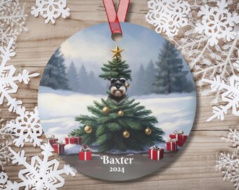 May include: A round Christmas ornament featuring a dog peeking out from behind a decorated Christmas tree. The dog is a miniature schnauzer with a gray and white coat. The ornament is decorated with a snowy winter scene and the text "Baxter 2024".