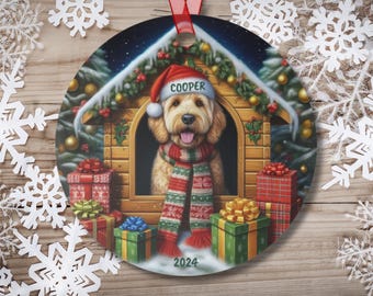 May include: A round Christmas ornament featuring a dog wearing a Santa hat and scarf, sitting in a wooden doghouse with presents and a snowy background. The dog's name, "Cooper", is written on the hat. The ornament also includes the year "2024".