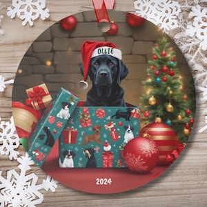 May include: A black Labrador Retriever wearing a Santa hat is inside a Christmas gift box with a green and red patterned design. The dog's name is "Ollie" and the box is surrounded by other Christmas gifts and ornaments. The year 2024 is printed on the bottom of the ornament.