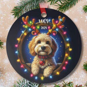 May include: A round Christmas ornament featuring a white dog with brown ears wearing a red collar with a bell. The dog is surrounded by a wreath of twinkling lights and has reindeer antlers with lights on them. The ornament has the text "Lucy 2024" on it.