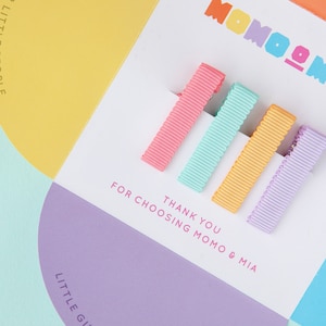Puede incluir: Un conjunto de cinco pinzas para el pelo de colores con un mensaje de agradecimiento que dice "Gracias por elegir Momo & Mia". Las pinzas para el pelo son en tonos de rosa, azul claro, naranja y morado.