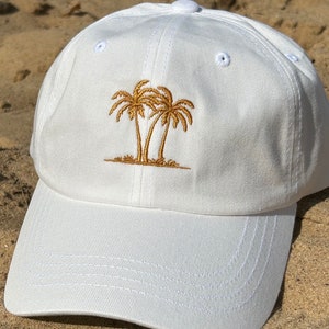 Könnte beinhalten: Weiße Baseballkappe mit einem goldenen gestickten Design aus drei Palmen.