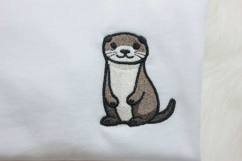 Op de afbeelding: Geborduurde afbeelding van een bruine en witte otter op een wit T-shirt.