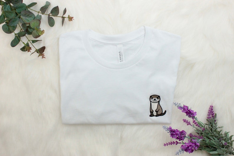 Op de afbeelding: Wit T-shirt met een geborduurd otterontwerp. De otter is bruin en zit rechtop.