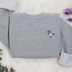 A sport grey unisex crew neck sweatshirt made of 50% cotton and 50% polyester is featured. It has a classic, soft, and warm fit, with a small embroidery on the left chest. The embroidery shows a small mallard duck standing and looking to the side.
