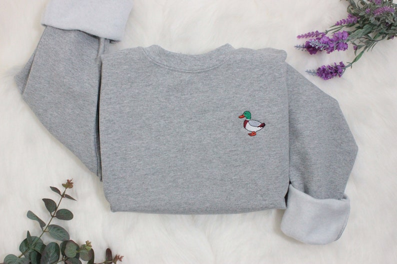 A sport grey unisex crew neck sweatshirt made of 50% cotton and 50% polyester is featured. It has a classic, soft, and warm fit, with a small embroidery on the left chest. The embroidery shows a small mallard duck standing and looking to the side.