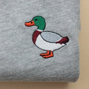 A sport grey unisex crew neck sweatshirt made of 50% cotton and 50% polyester is featured. It has a classic, soft, and warm fit, with a small embroidery on the left chest. The embroidery shows a small mallard duck standing and looking to the side.