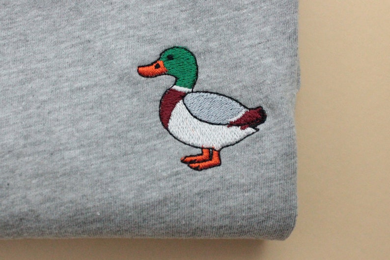 A sport grey unisex crew neck sweatshirt made of 50% cotton and 50% polyester is featured. It has a classic, soft, and warm fit, with a small embroidery on the left chest. The embroidery shows a small mallard duck standing and looking to the side.