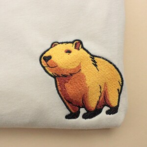Short-sleeved soft cream T-shirt with a crew neckline, made from 100% combed and ring-spun cotton, features a small capybara embroidery on the left chest. The embroidery shows a standing small cartoon-style capybara capybara smiling, looking aside.