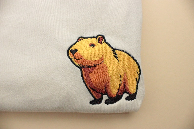 Short-sleeved soft cream T-shirt with a crew neckline, made from 100% combed and ring-spun cotton, features a small capybara embroidery on the left chest. The embroidery shows a standing small cartoon-style capybara capybara smiling, looking aside.