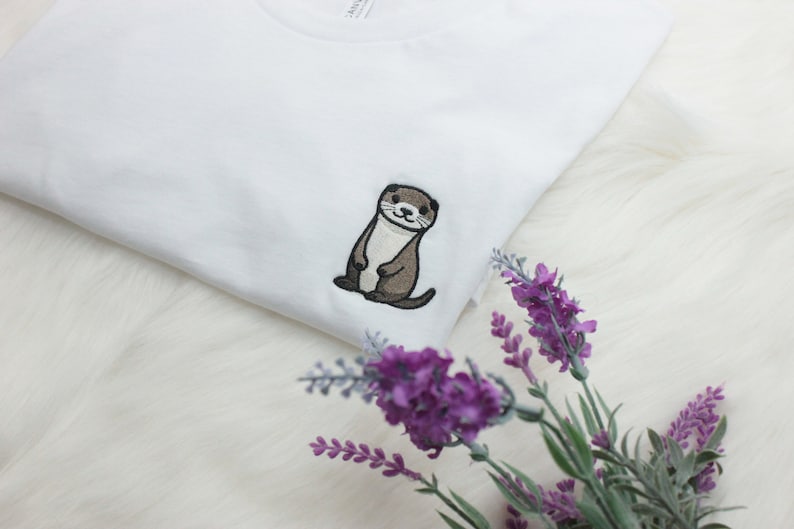 Op de afbeelding: Wit T-shirt met een geborduurde bruine otter op de voorkant. De otter staat rechtop met zijn poten voor zich. Het T-shirt ligt op een wit, pluche oppervlak met paarse bloemen.