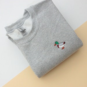 A sport grey unisex crew neck sweatshirt made of 50% cotton and 50% polyester is featured. It has a classic, soft, and warm fit, with a small embroidery on the left chest. The embroidery shows a small mallard duck standing and looking to the side.