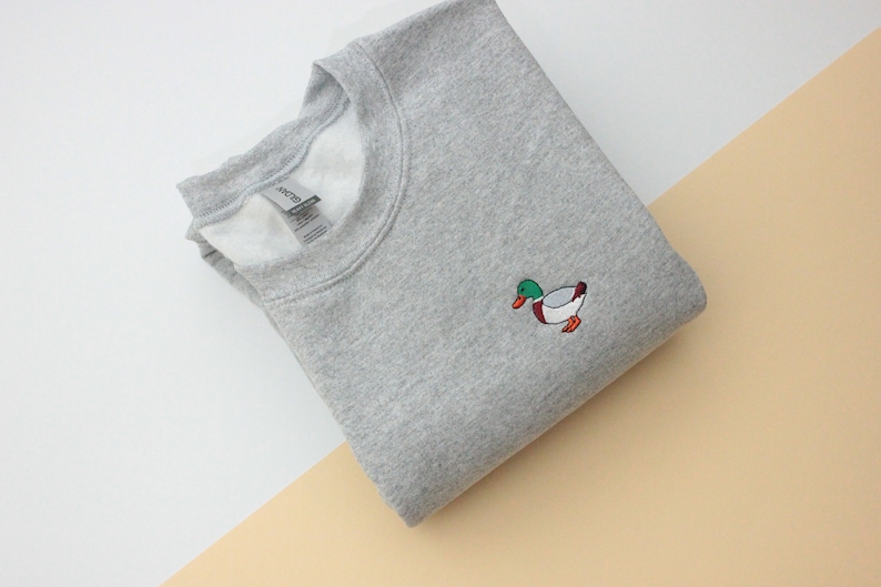 A sport grey unisex crew neck sweatshirt made of 50% cotton and 50% polyester is featured. It has a classic, soft, and warm fit, with a small embroidery on the left chest. The embroidery shows a small mallard duck standing and looking to the side.