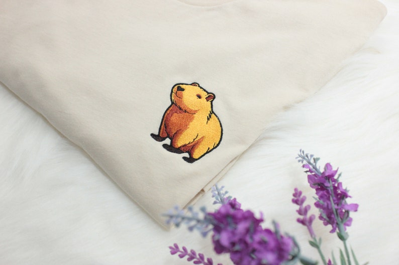 Short-sleeved soft cream T-shirt with a crew neckline, made from 100% combed and ring-spun cotton, features a small capybara embroidery on the left chest. The embroidery shows a standing small cartoon-style capybara capybara smiling, looking aside.