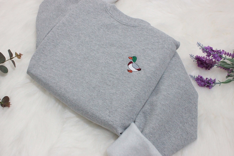 A sport grey unisex crew neck sweatshirt made of 50% cotton and 50% polyester is featured. It has a classic, soft, and warm fit, with a small embroidery on the left chest. The embroidery shows a small mallard duck standing and looking to the side.