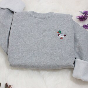 A sport grey unisex crew neck sweatshirt made of 50% cotton and 50% polyester is featured. It has a classic, soft, and warm fit, with a small embroidery on the left chest. The embroidery shows a small mallard duck standing and looking to the side.