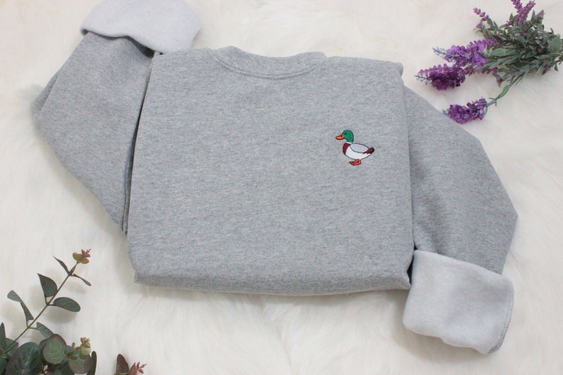 A sport grey unisex crew neck sweatshirt made of 50% cotton and 50% polyester is featured. It has a classic, soft, and warm fit, with a small embroidery on the left chest. The embroidery shows a small mallard duck standing and looking to the side.
