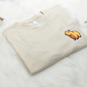 Short-sleeved soft cream T-shirt with a crew neckline, made from 100% combed and ring-spun cotton, features a small capybara embroidery on the left chest. The embroidery shows a standing small cartoon-style capybara capybara smiling, looking aside.