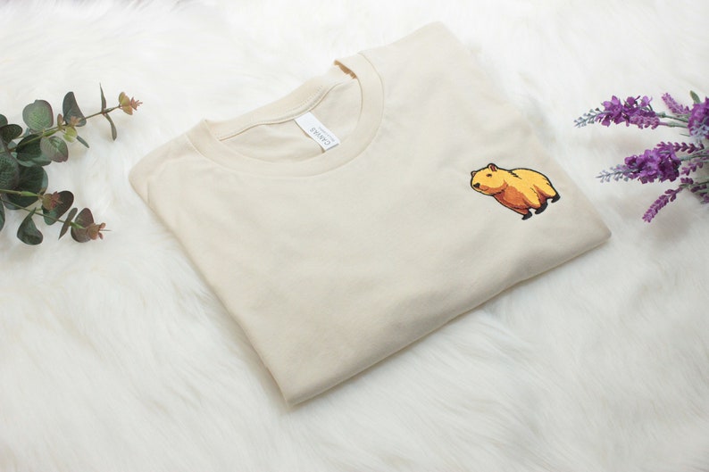 Short-sleeved soft cream T-shirt with a crew neckline, made from 100% combed and ring-spun cotton, features a small capybara embroidery on the left chest. The embroidery shows a standing small cartoon-style capybara capybara smiling, looking aside.