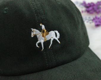 May include: A dark green corduroy baseball cap with a white embroidered image of a person riding a horse.