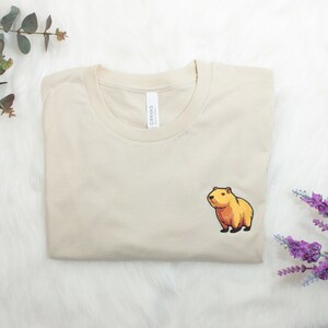 Short-sleeved soft cream T-shirt with a crew neckline, made from 100% combed and ring-spun cotton, features a small capybara embroidery on the left chest. The embroidery shows a standing small cartoon-style capybara capybara smiling, looking aside.