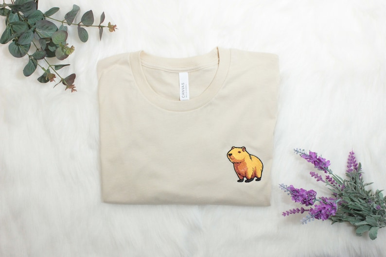 Short-sleeved soft cream T-shirt with a crew neckline, made from 100% combed and ring-spun cotton, features a small capybara embroidery on the left chest. The embroidery shows a standing small cartoon-style capybara capybara smiling, looking aside.