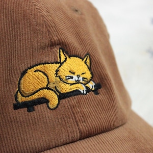 Peut inclure: Casquette de baseball en velours côtelé marron avec un chat jaune endormi brodé sur une surface noire. Le chat a des détails blancs et est dans une pose détendue. La casquette a une visière incurvée et un design classique à six panneaux.