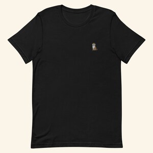 A short-sleeved black T-shirt with a crew neckline, made from 100% combed and ring-spun cotton, features a small otter embroidery on the left chest. The embroidery shows a standing cartoon-style otter tucking its paws and smiling sweetly.