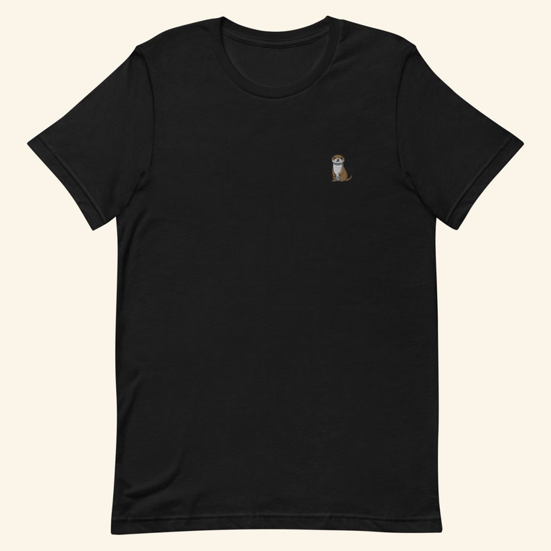 A short-sleeved black T-shirt with a crew neckline, made from 100% combed and ring-spun cotton, features a small otter embroidery on the left chest. The embroidery shows a standing cartoon-style otter tucking its paws and smiling sweetly.