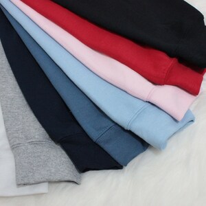 Op de afbeelding: Een stapel van acht verschillende gekleurde sweatshirts, waaronder wit, grijs, donkerblauw, denimblauw, lichtblauw, roze, rood en zwart.