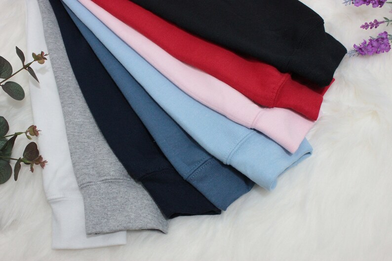 Op de afbeelding: Een stapel van acht verschillende gekleurde sweatshirts, waaronder wit, grijs, donkerblauw, denimblauw, lichtblauw, roze, rood en zwart.