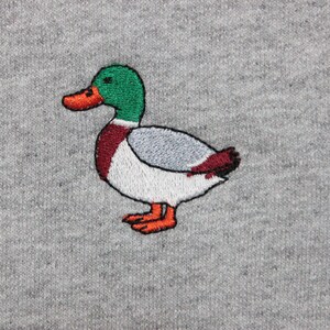 A sport grey unisex crew neck sweatshirt made of 50% cotton and 50% polyester is featured. It has a classic, soft, and warm fit, with a small embroidery on the left chest. The embroidery shows a small mallard duck standing and looking to the side.