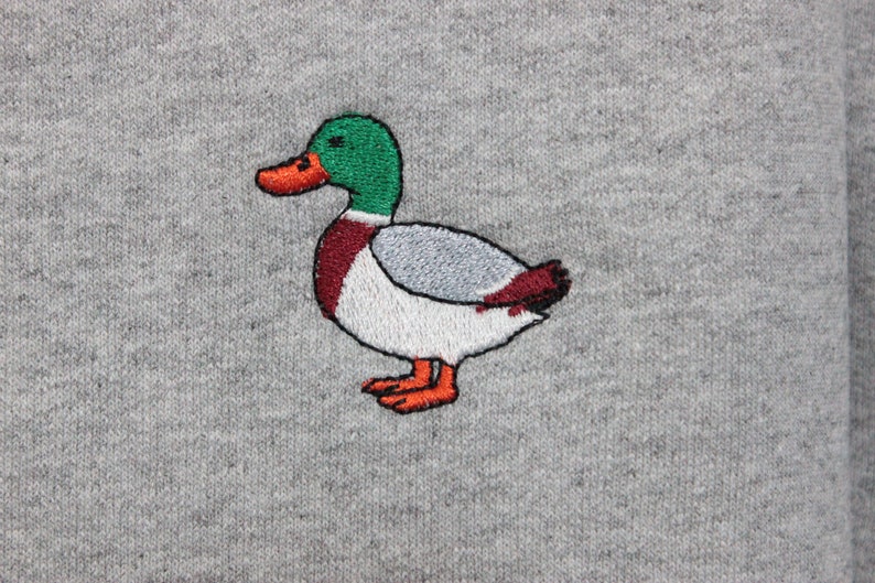 A sport grey unisex crew neck sweatshirt made of 50% cotton and 50% polyester is featured. It has a classic, soft, and warm fit, with a small embroidery on the left chest. The embroidery shows a small mallard duck standing and looking to the side.